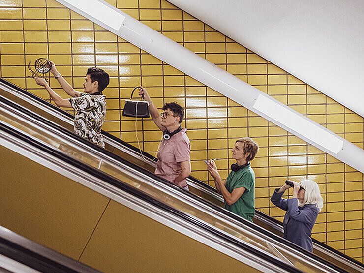 Die Veranstaltre*innen des Musikbingos fahren eine Rolltreppe hoch und machen Faxen. Gelbe Kacheln im Hintergrund.