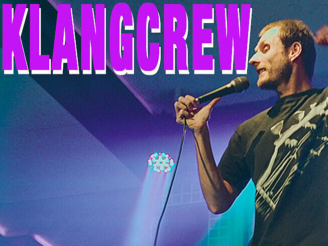 Der Chorleiter Nick "Chillionair" Hämmerling rappt auf der Bühne. "Klangcrew" steht in Pink oben links.