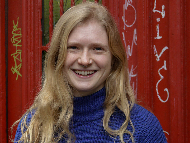 Nele hat lange blonde Haare, steht vor dem Eigang zur OKJA und lächelt in die Kamera.