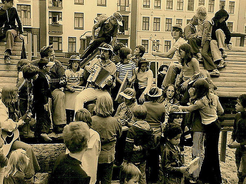 Ein schwar-weiß Foto zeigt Besucher*innen des Sommerfests von 1976  auf dem Hein-Köllisch-Platz. Ein Mann spielt Gitarre und einerAkkordeon.