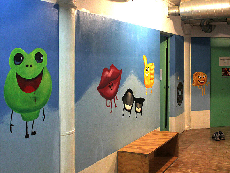 Eine bunt angemalte Wand aus dem Kinderbereich. Ein Frosch, ein Mund, ein hochgestreckter Daumen auf blauem Grund,