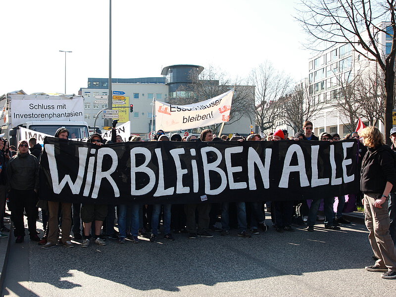 Auf einer Demo für die Esso Häuser. Ein großes schwarzes Transpartent mit weißer Schrift wird gehalten: Wir bleiben Alle! 