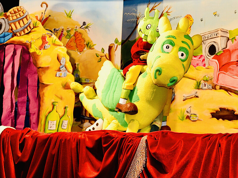 Puppenspiel: Ein Olchi reitet auf gelb-grünem Drache vor der Kulisse des Müllbergs