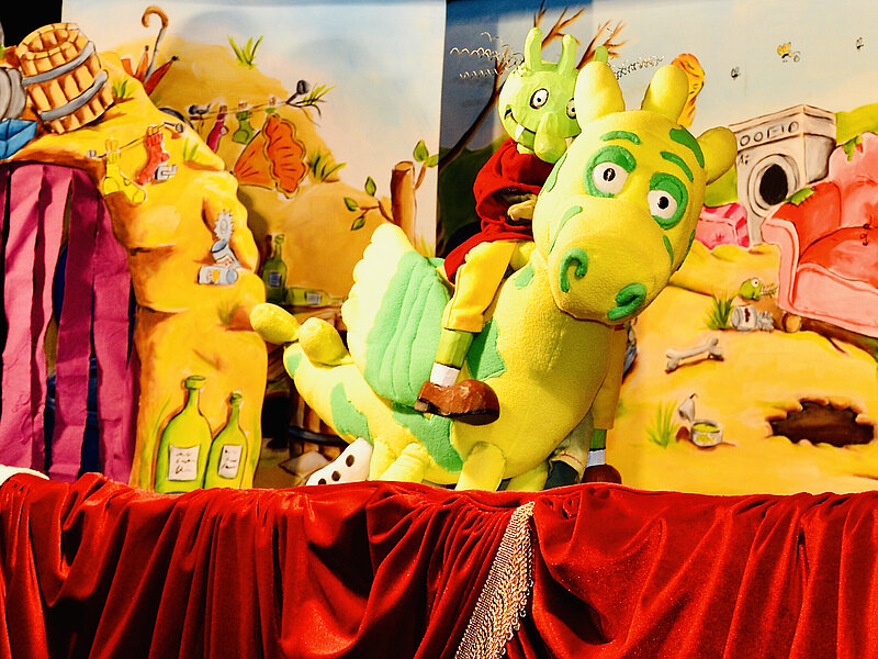 Puppenspiel: Ein Olchi reitet auf gelb-grünem Drache vor der Kulisse des Müllbergs