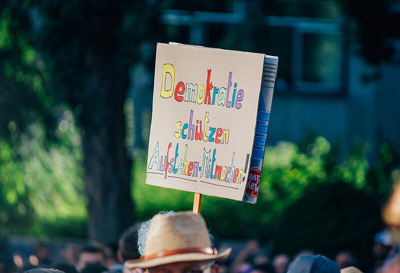 Ein Demoschild "Demokratie schützen - Aufstehen - mitmachen" in bunten Buchstaben. Von der Person, die es trägt ist nur ein Strohhut zu sehen.