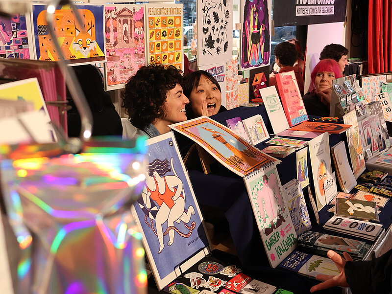 Fröhliche Personen sitzen an einem Messestand zwischen bunten Comics und Drucken.