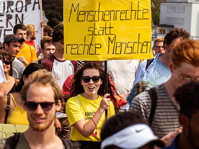 Auf einer Demo hält eine Frau mit gelbem T-Shirt von amnesty international ein gelbes Schild hoch "Menschenrechte statt rechte Menschen".