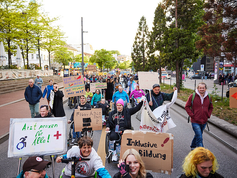 Foto von einer Demo zum 5. Mai, dem Europäischen Protesttag zur Gleichstellung von Menschen mit Behinderung. Eine bunte Menge mit Schildern, teilweise Rollifahrenden.
