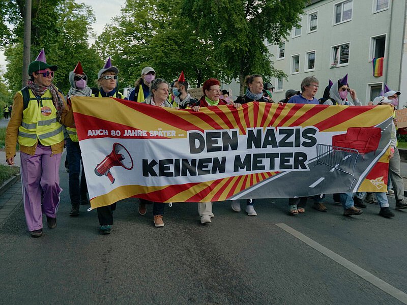 8 Demonstrierende halten vor sich ein großes Transparent "Auch 80 Jahre später ... den Nazis keinen Meter". Auf dem Transpi ist außerdem eine Straße abgebildet, die mit einem Absperrgitter blockiert ist. Dahintr steht ein rotes Sofa. Die Demonstrierenden sind unterschiedlichen Alters und haben teilweise pinke Maskten auf.