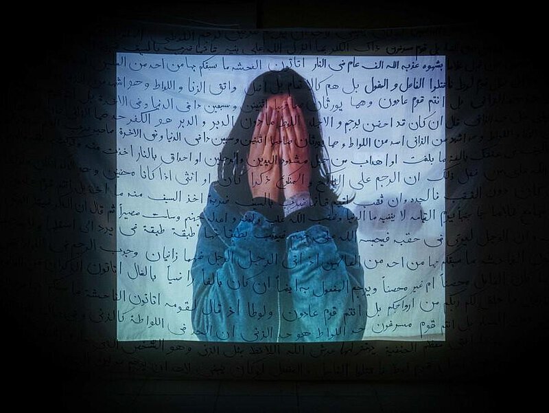 Foto einer Person, die sich die Hände vor das GEsicht hält. Darauf projizierte arabische Schriftzeichen.