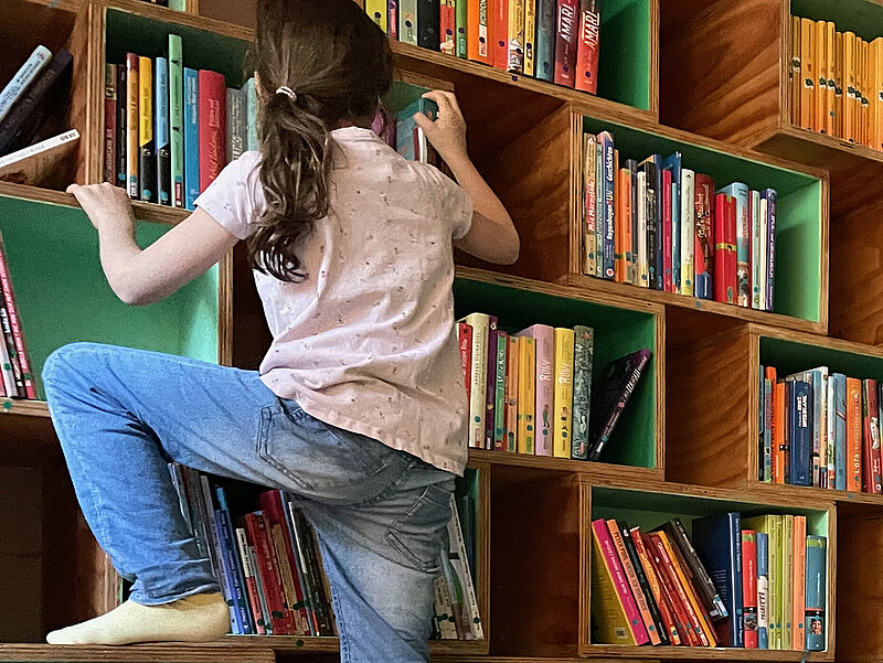 Ein Kind mit Pferdeschwanz klettert die Bücherregal-Wand hoch.