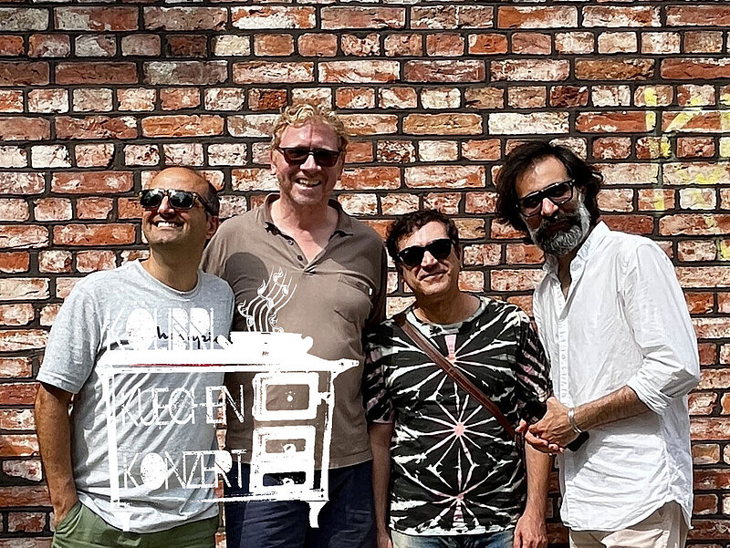 Bandfoto: 4 Männer stehen vor einer Backsteinwand und lachen in die Kamera