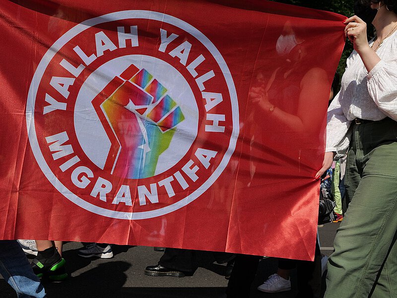 Auf einer Demo wird die Fahne der Migrantifa getragen. Rote Fahne, bunte Faust und der Slogan "Yallah, Yallah, Migrantifa"